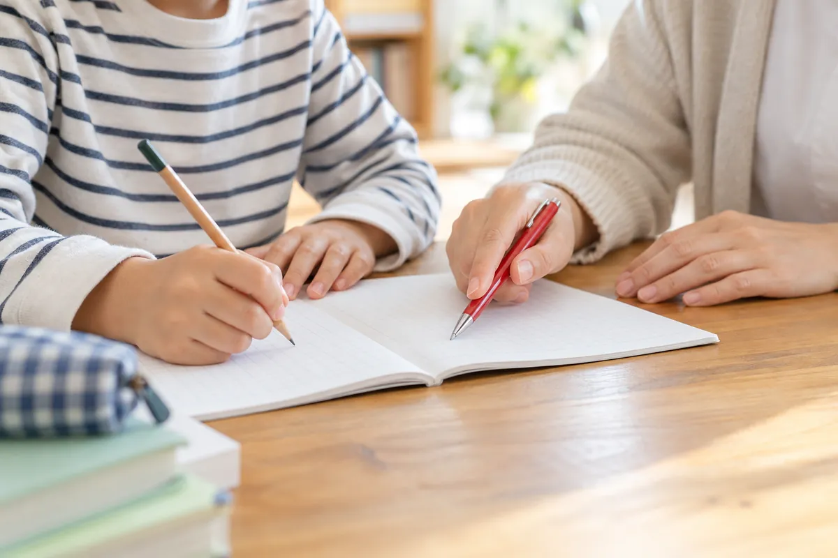 ノートに書き込む子どもの手元と、赤いペンを持って寄り添う大人の手